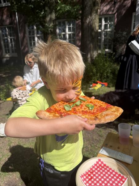 A young child with tiger face paint joyfully eats a slice of pizza topped with basil, surrounded by green trees and an old brick building